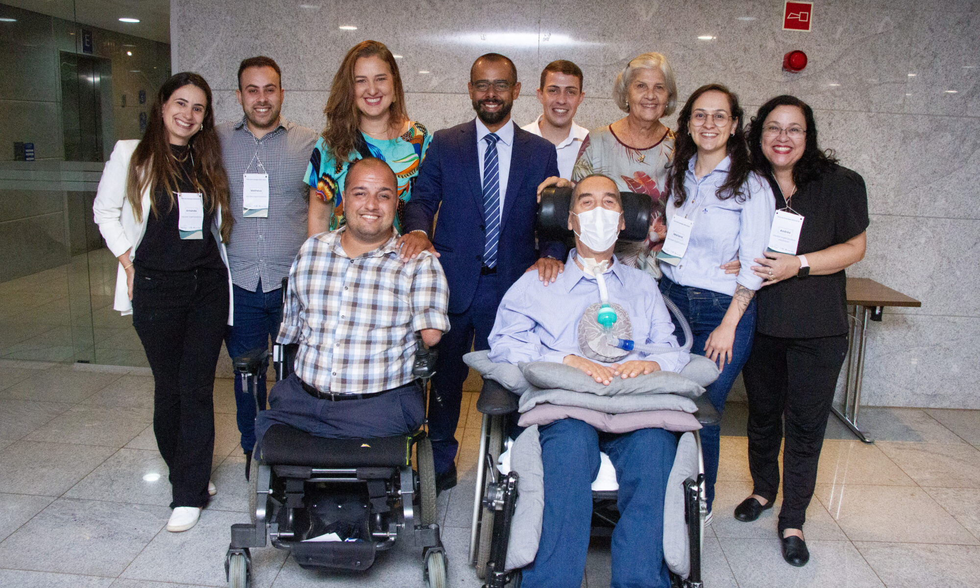 Um grupo de pessoas constituído por 5 mulheres e 5 homens sorrindo. Dois homens estão em cadeiras de rodas. Os Asiders Matheus Garcia e Mariana Oliveira estão neste grupo.
