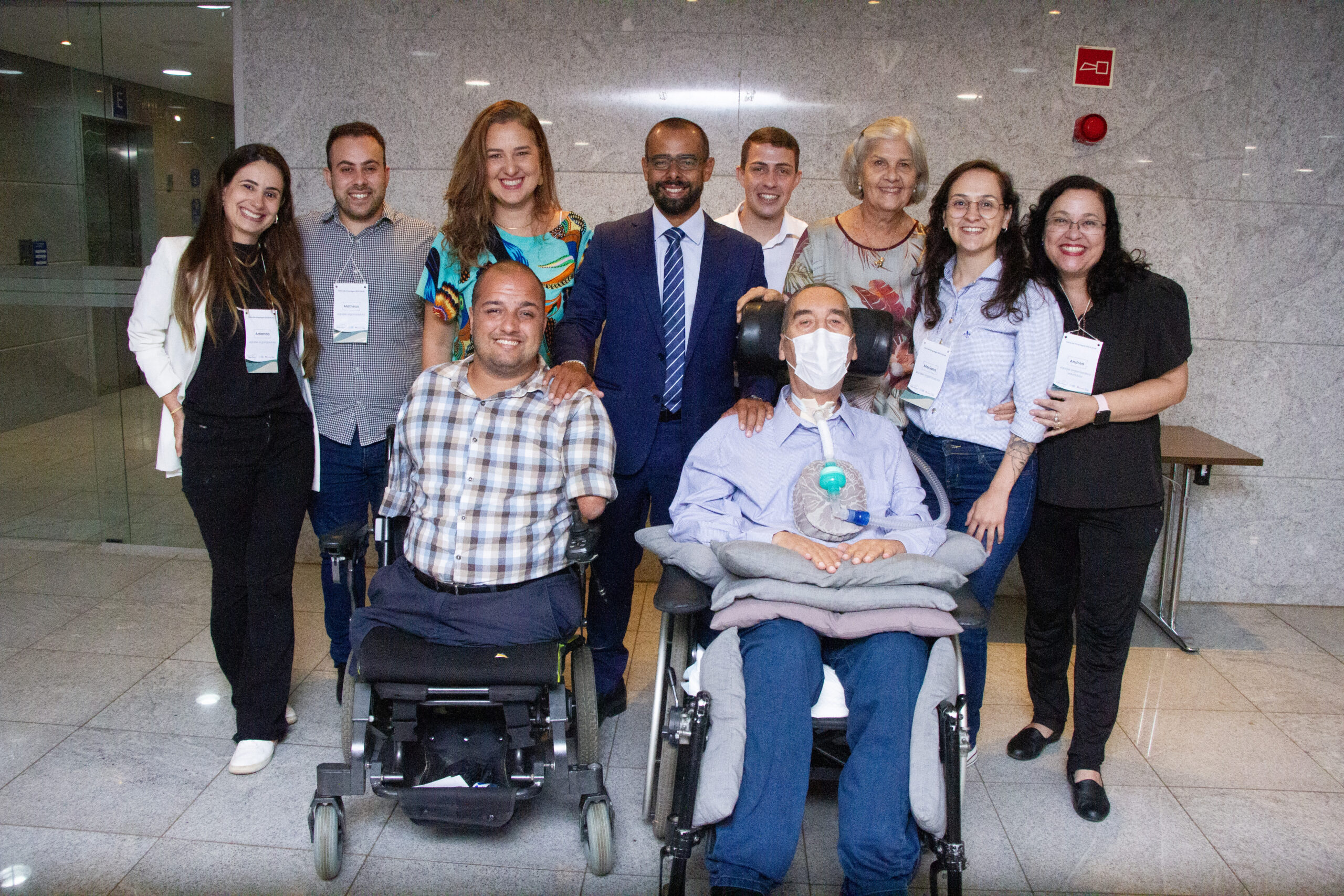 Um grupo de pessoas constituído por 5 mulheres e 5 homens sorrindo. Dois homens estão em cadeiras de rodas. Os Asiders Matheus Garcia e Mariana Oliveira estão neste grupo.