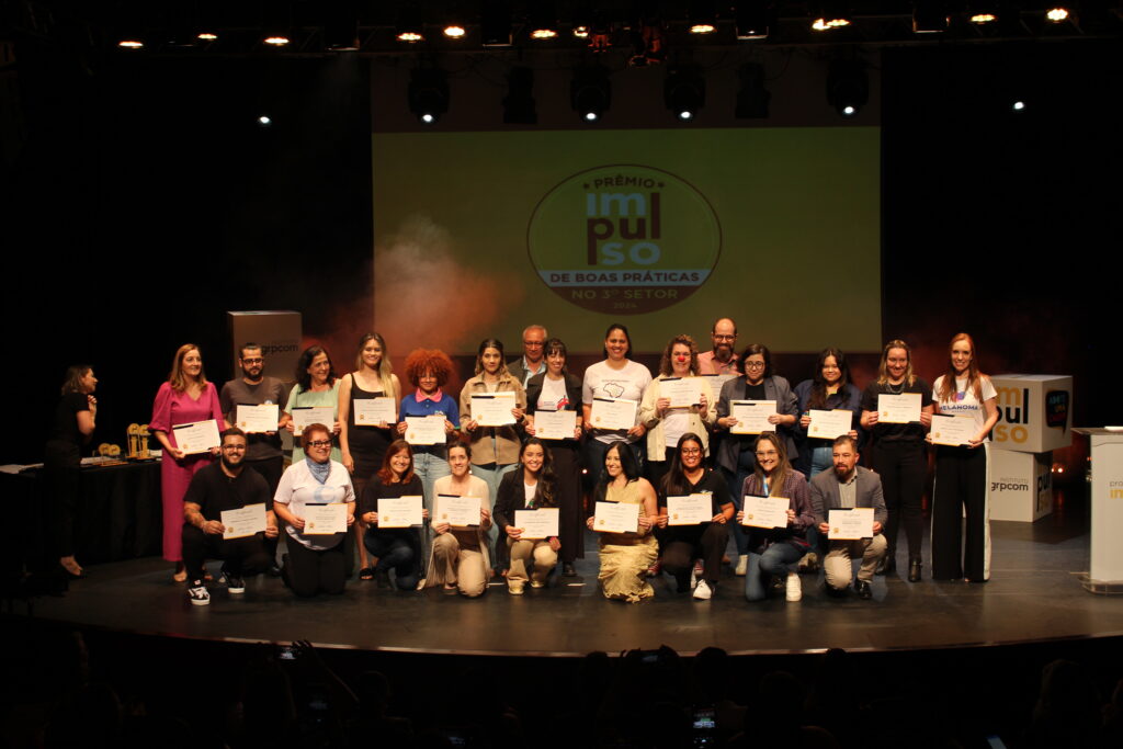 Pessoas no palco da cerimônia do Prêmio Impulso em formação de time de futebol, segurando um certificado de reconhecimento.